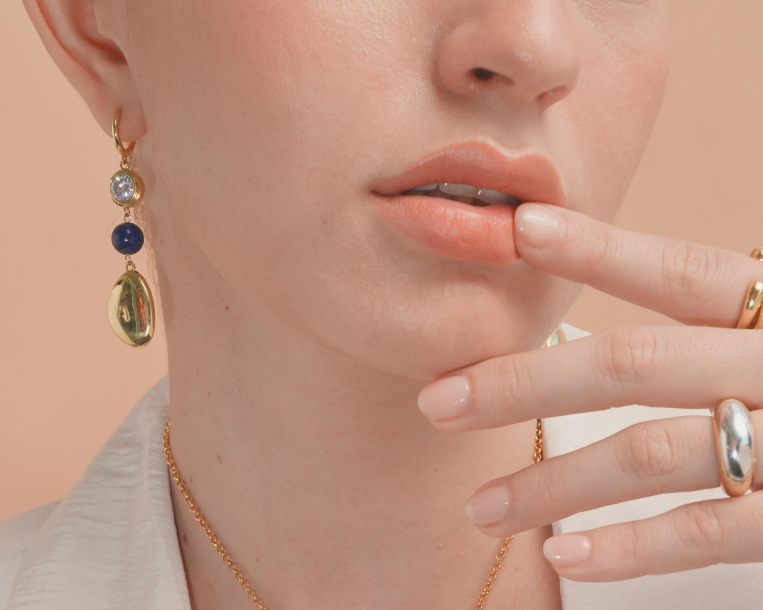 Close-up of a woman wearing a gold necklace with blue stones, earrings, and rings on a light pink background.
