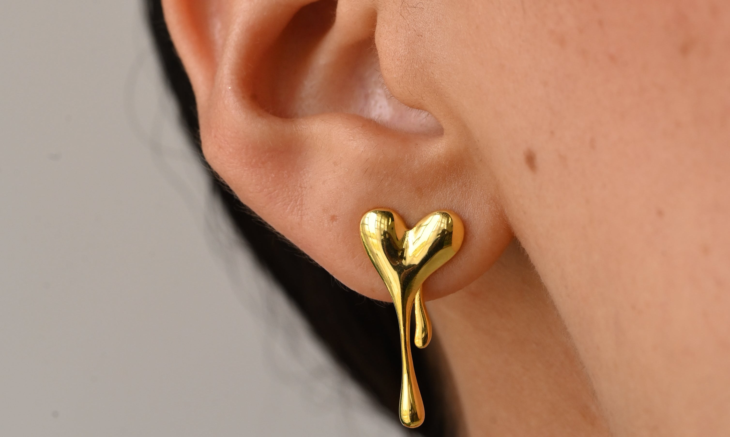 A close-up of a person's ear wearing a small gold heart-shaped drop earring against a plain light gray background.