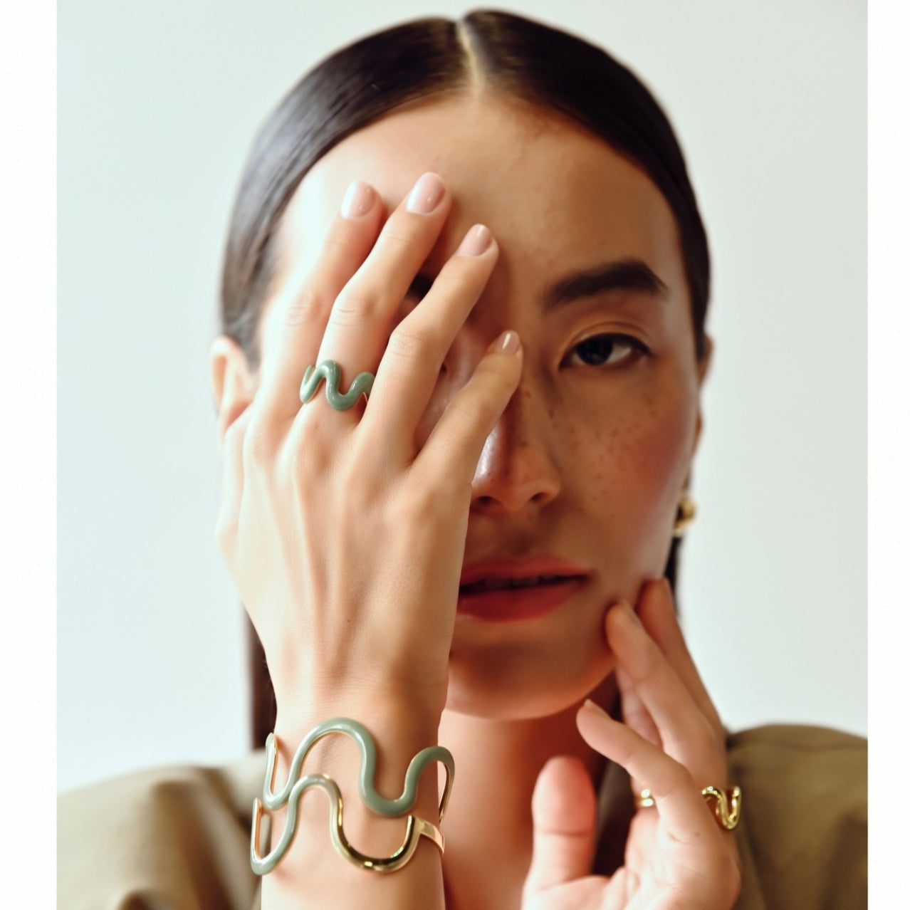 A close-up portrait of a woman wearing gold-toned jewelry, including a gold snake ring on one finger and a matching gold snake bracelet on her wrist, with a light olive green enamel accent. She is wearing a light olive green blazer against a clean, light gray background.