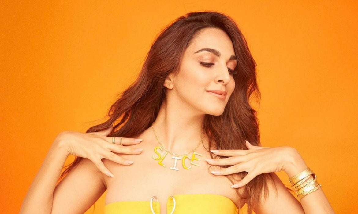 A woman posing against a vibrant orange background, wearing a matching yellow strapless tube top and skirt set with a silver zipper detail at the bust, accessorized with a yellow and white necklace and gold bracelets.