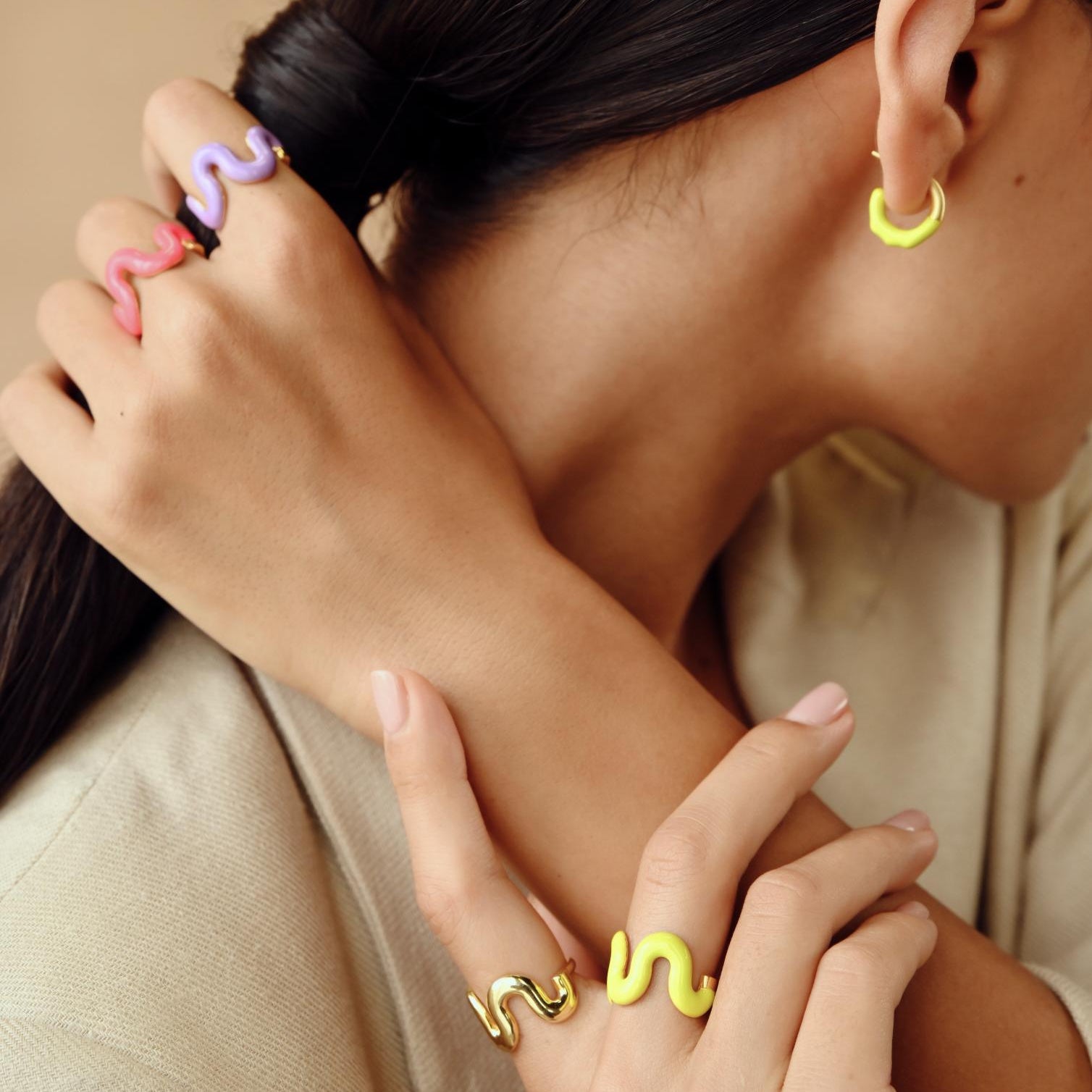 A close-up lifestyle shot of a model wearing colorful snake-shaped rings on her fingers and a small yellow hoop earring, dressed in a beige blazer against a warm beige background.