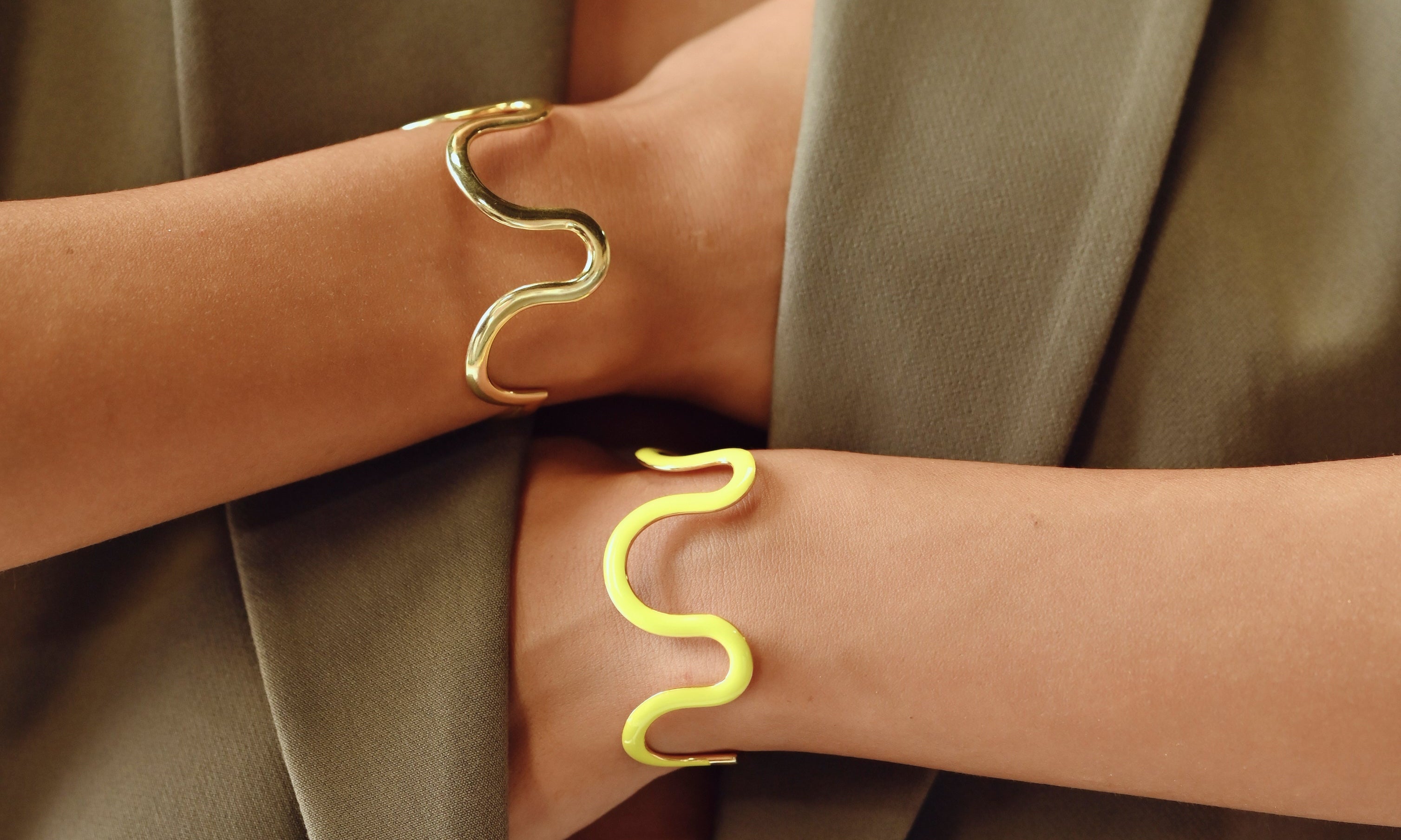 Two gold snake-shaped cuff bracelets worn on crossed forearms, shown close-up against an olive green blazer.