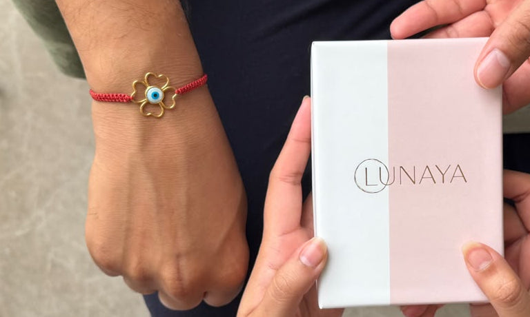 A close-up lifestyle shot of a person receiving a pink and white Lunaya jewelry box from another person. The person holding the box is wearing a gold bracelet, while the person giving the box is wearing a red string bracelet with a gold flower charm containing a blue and green evil eye.